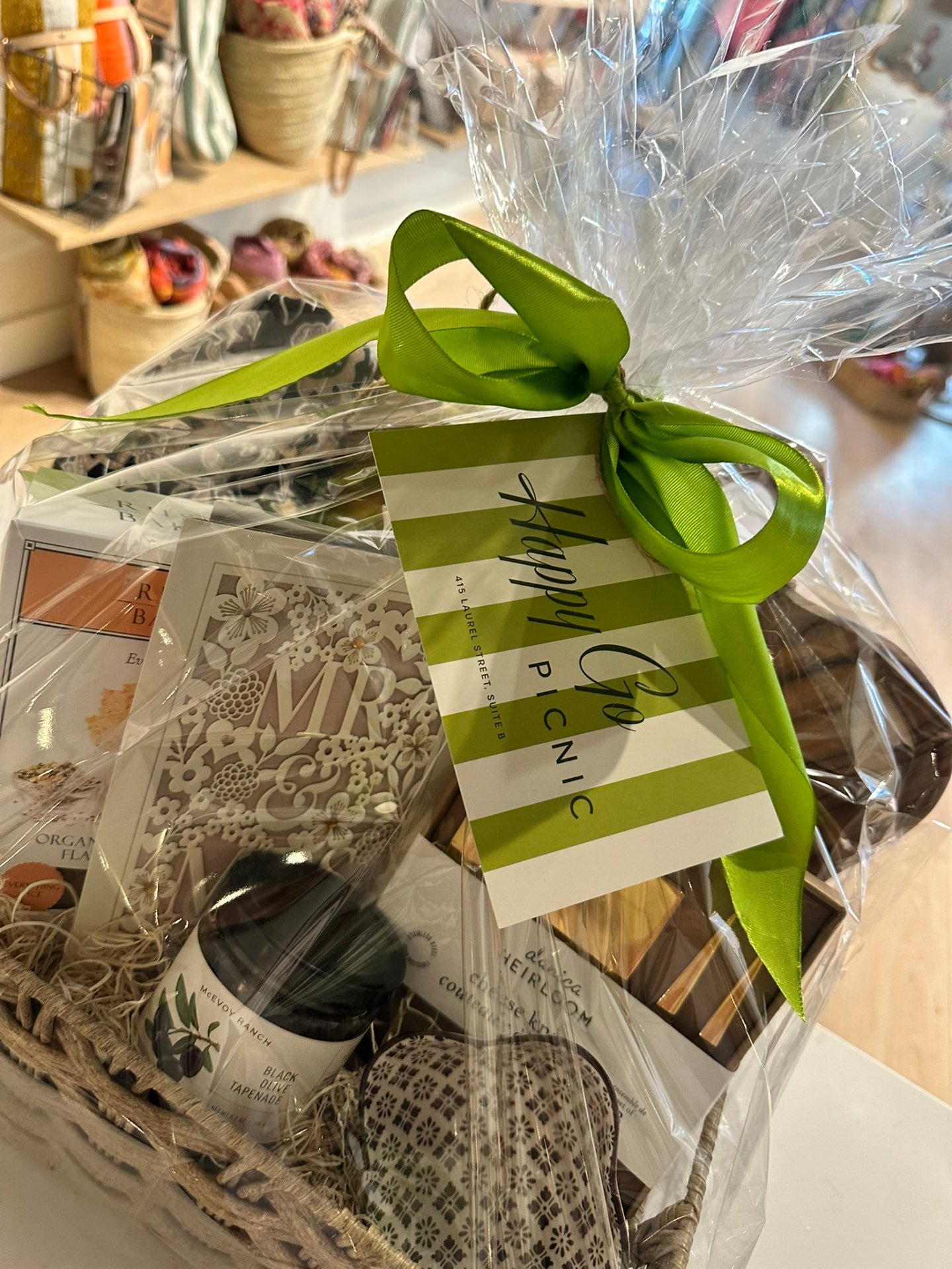 A gift basket wrapped in cellophane with a green ribbon and striped Happy Go Picnic tag