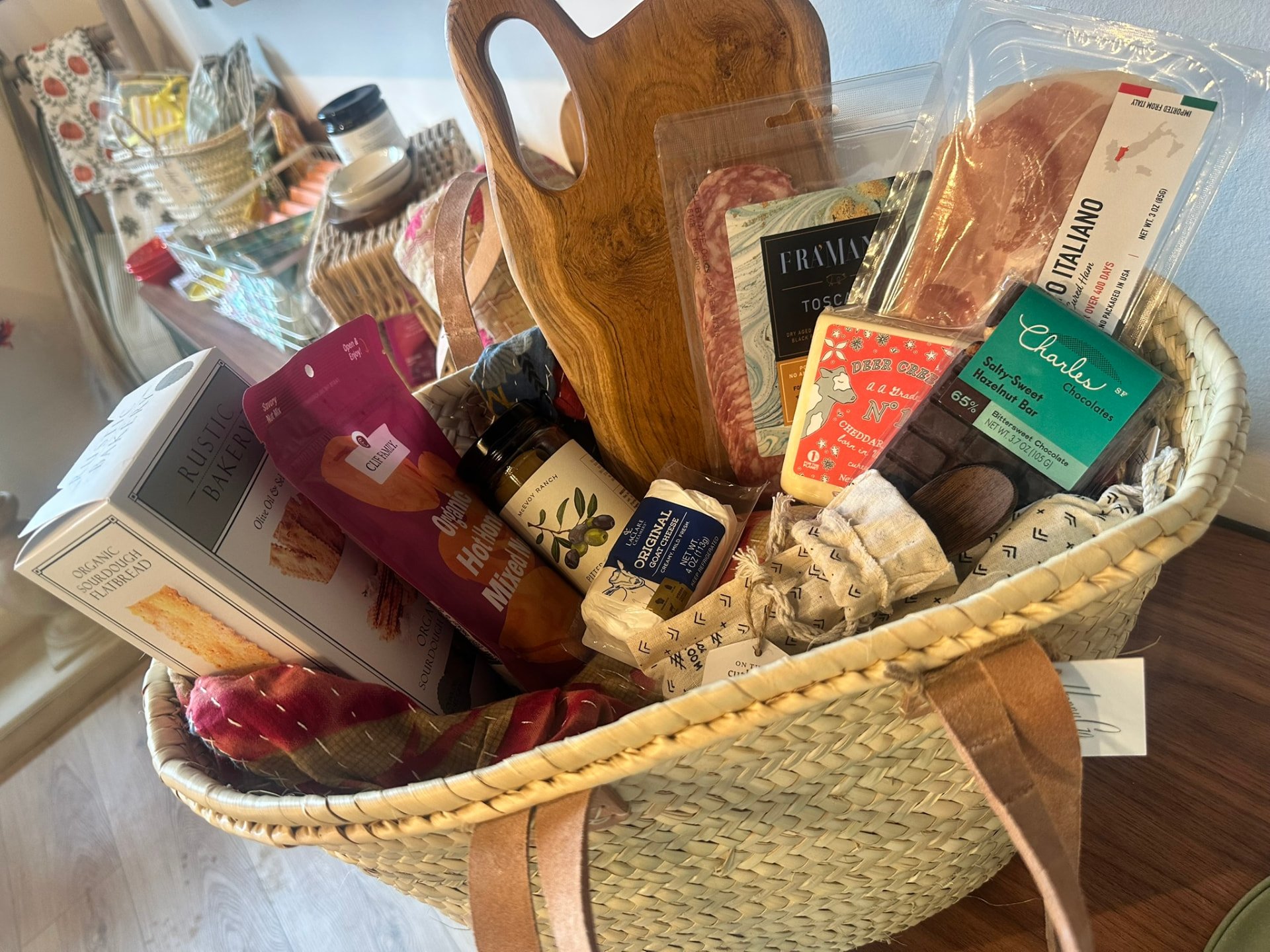 A basket filled with crackers, salami, olive oil, chocolate and an olive-wood board