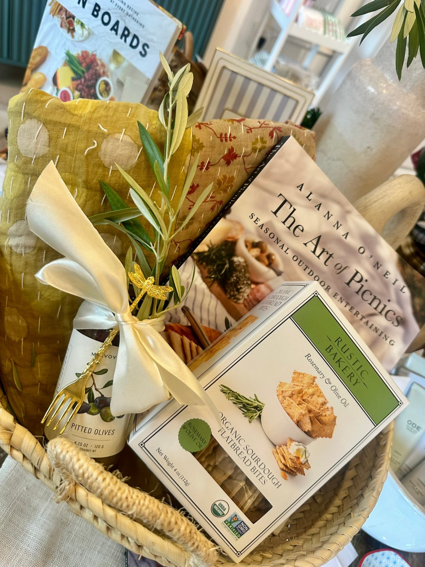 A woven gift basket with olives tied in a cream ribbon, sourdough crackers and The Art of Picnics book