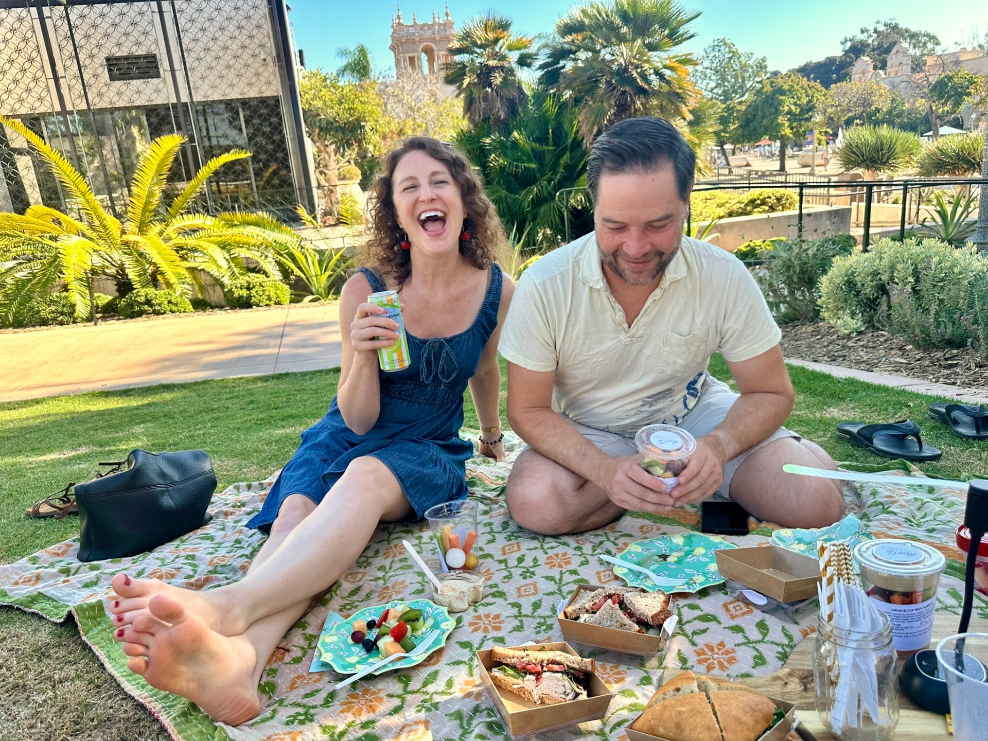 Two friends laughing on a picnic blanket with sandwiches and fruit near the Botanical Building
